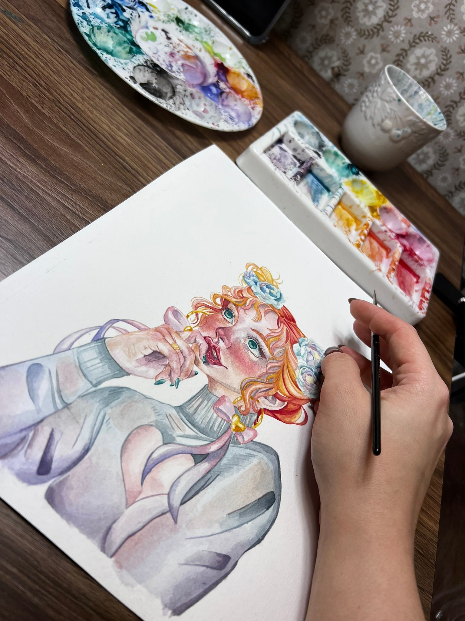 Photo of a watercolor painting work in progress. The artist is holding the paintbrush off to the side and you can see the pallets covered in paint and the glass for cleaning the brush.